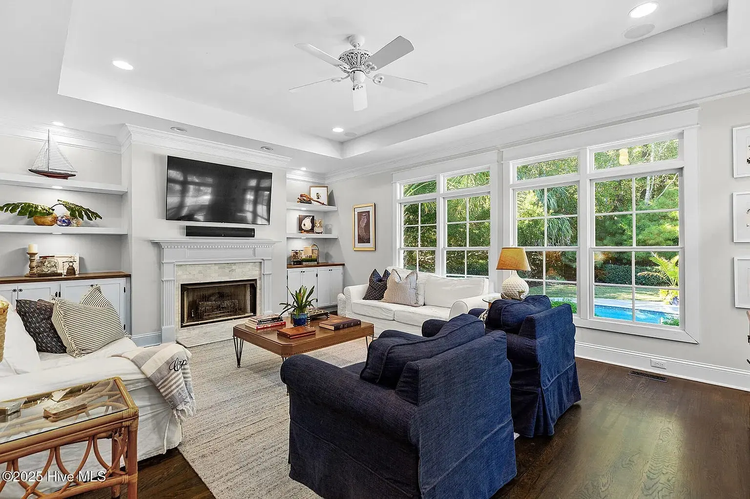Spacious living room with large windows, blue armchairs, white sofas, and a wall-mounted TV above a fireplace.