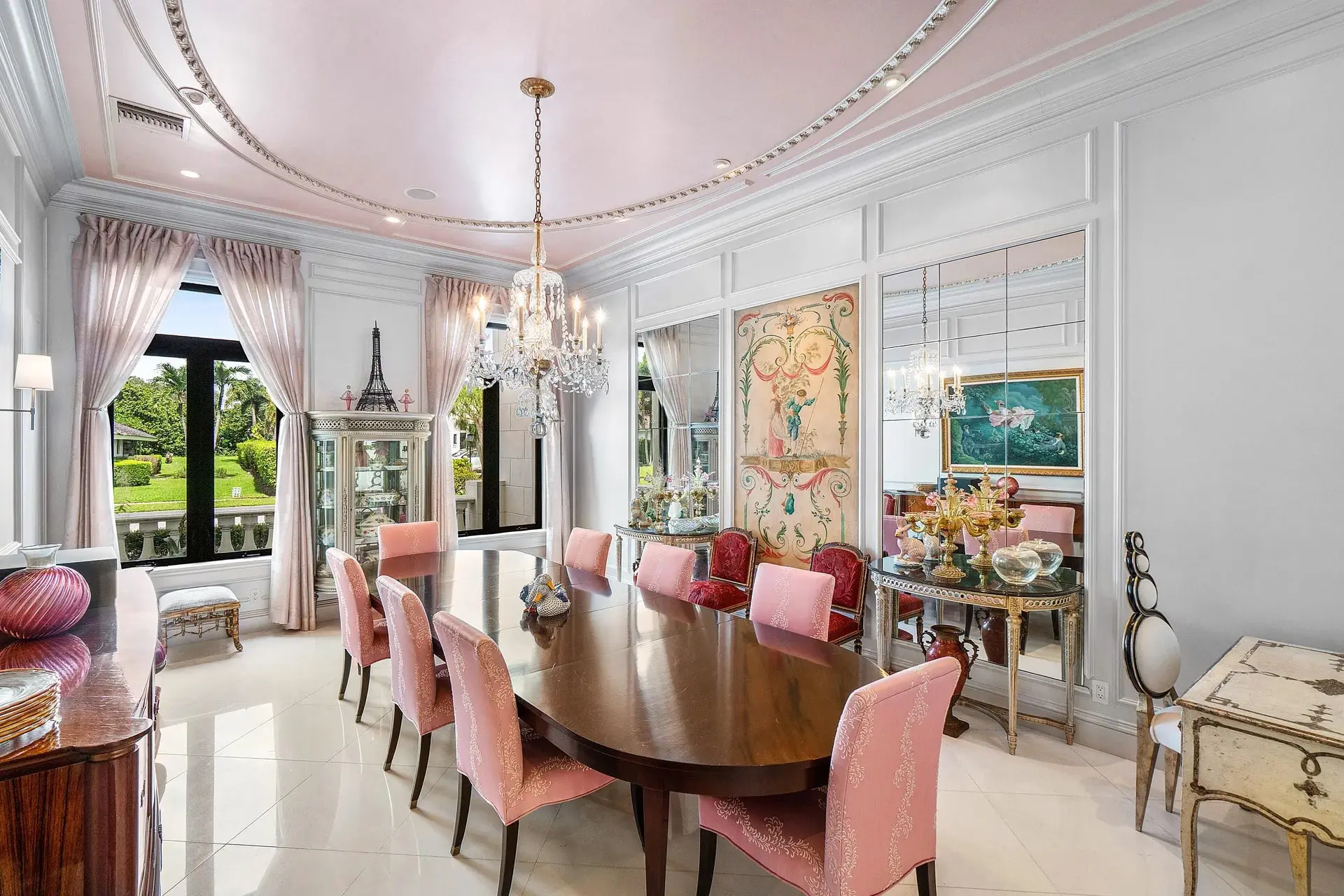 Elegant dining room with a long wooden table, pink chairs, chandeliers, and large windows.