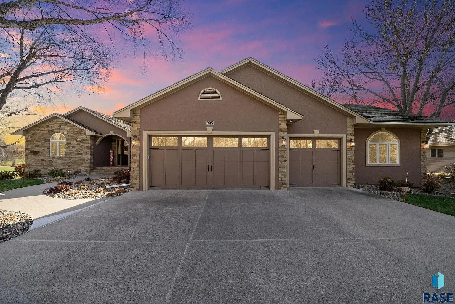 Single-story house with three-car garage, stone accents, and a sunset sky in the background.
