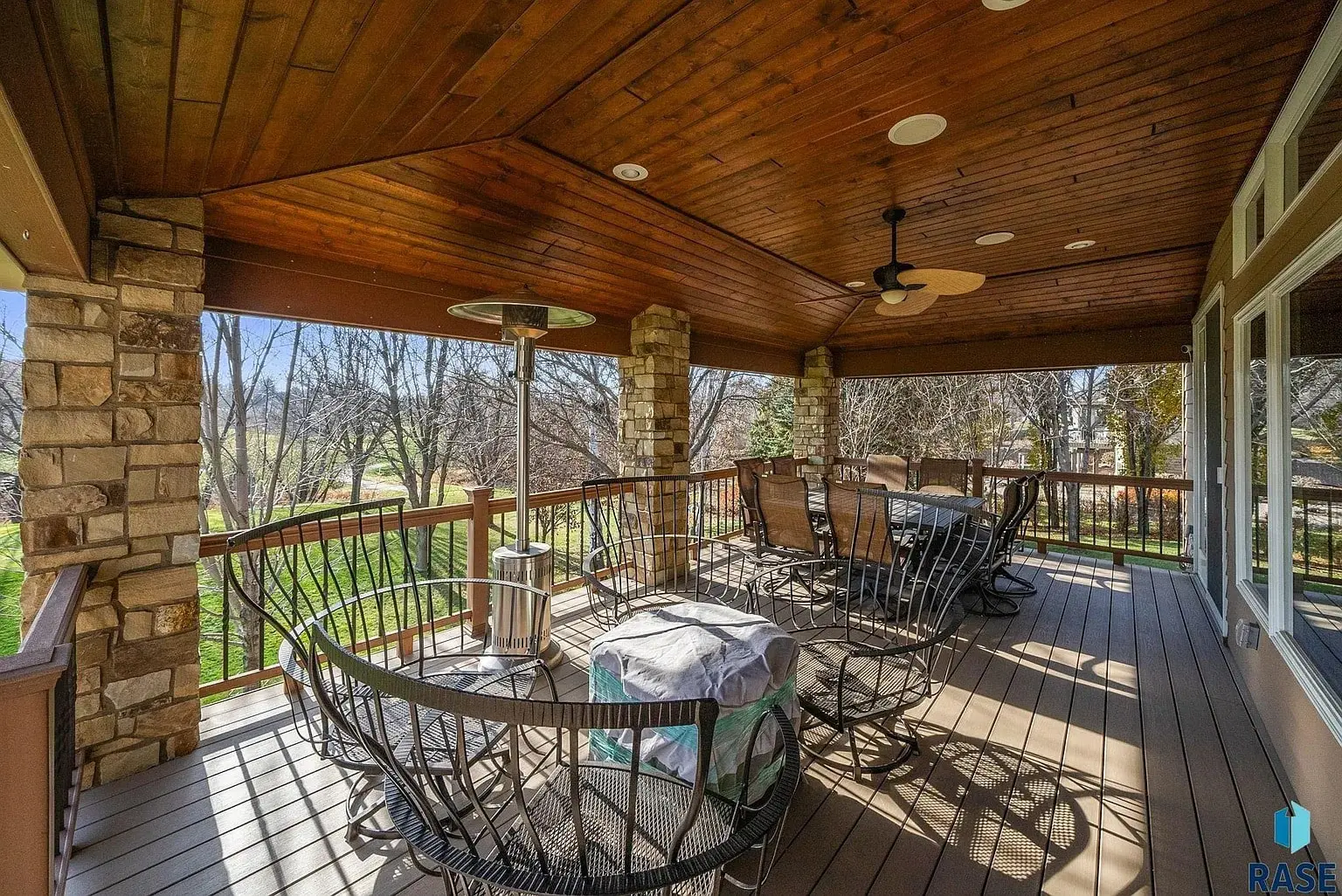 Covered wooden deck with dining table, chairs, ceiling fan, and view of trees and lawn.
