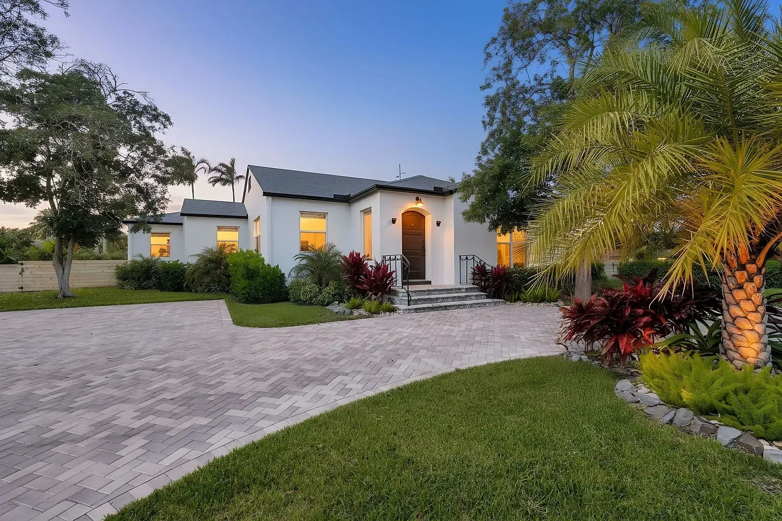 Modern white house with large windows, surrounded by lush greenery and a paved driveway at sunset.