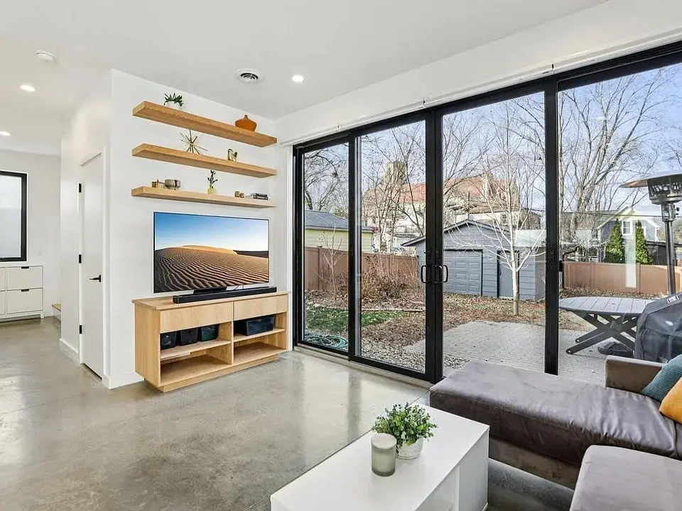 Modern living room with large glass doors, wall-mounted shelves, and a TV. Outdoor view of trees.