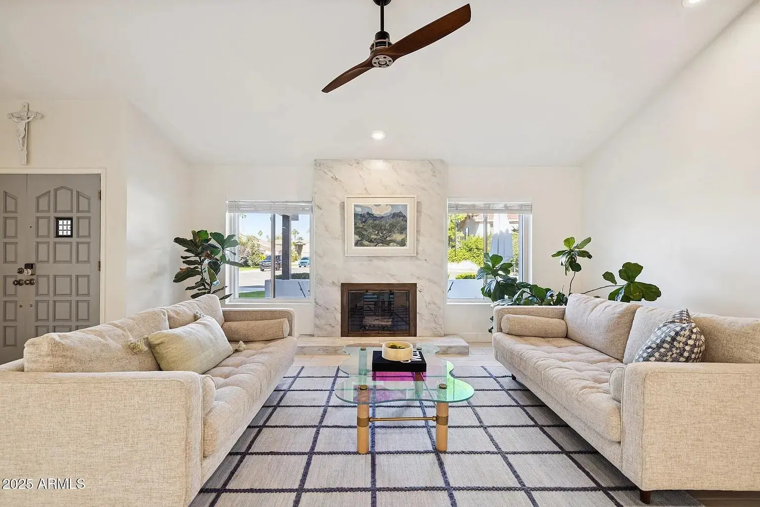 Living room with two beige sofas, glass coffee table, and fireplace, decorated with plants.