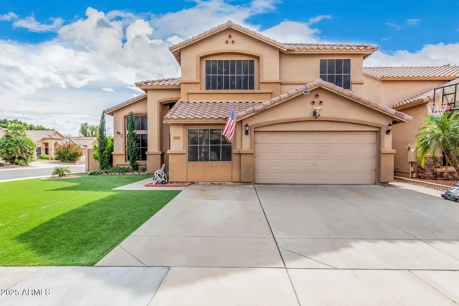 Two-story beige house with a manicured lawn, American flag, and a two-car garage.