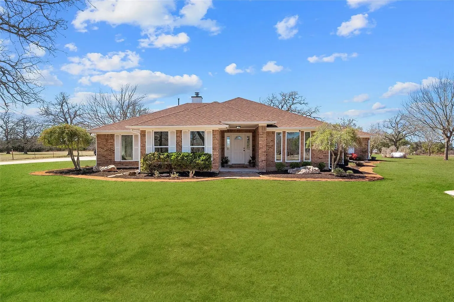 Single-story brick house with a brown roof, surrounded by a large green lawn and trees.