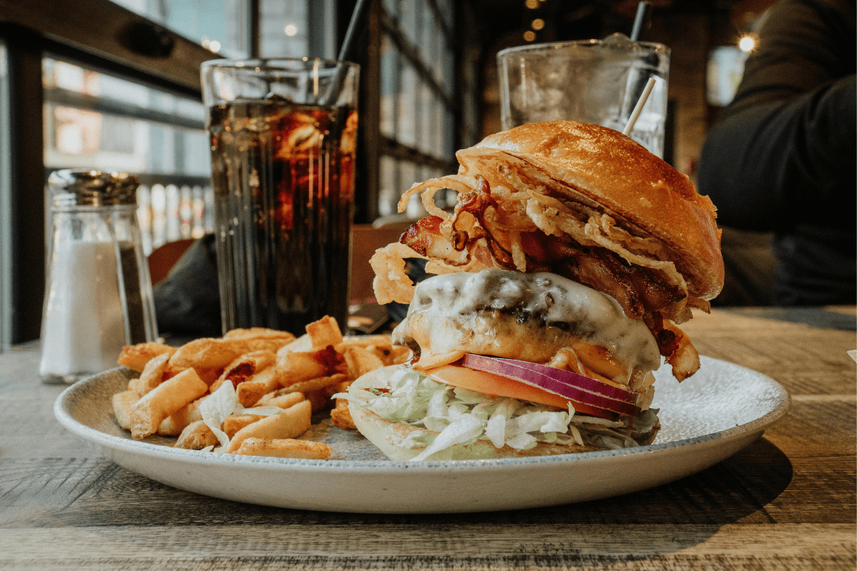 A fully stacked burger sitting on a plate with french fries and a drink behind it.