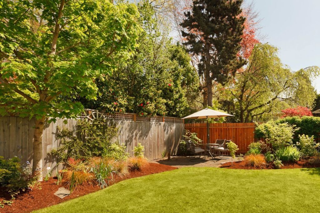 A sunny backyard with a green lawn, wood fence, and a small patio set with an umbrella surrounded by trees and shrubs.