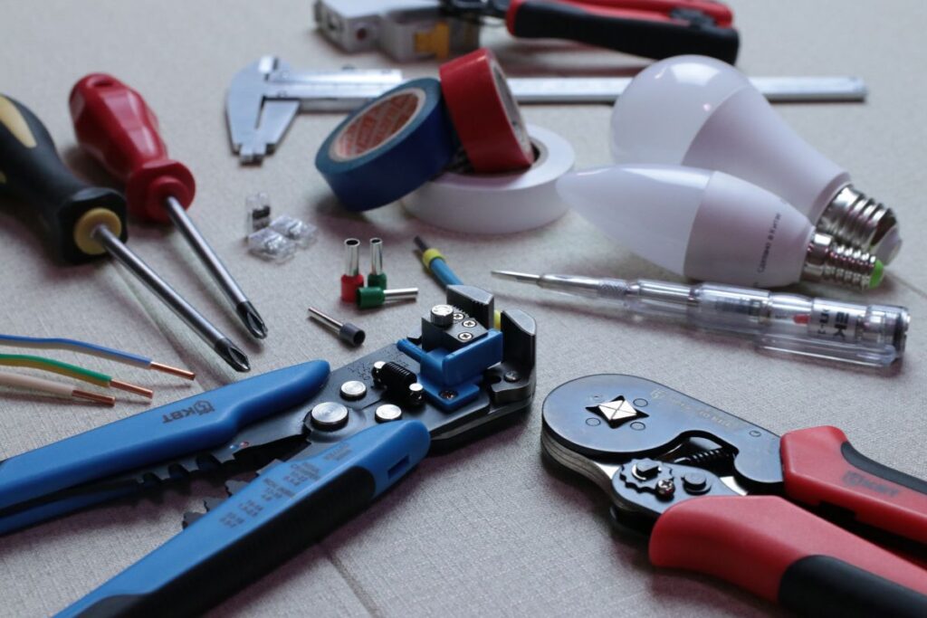 Electrical tools laid out on a surface: screwdrivers, wire strippers and crimpers, rolls of tape, stripped wires with connectors, a voltage tester, and LED bulbs.