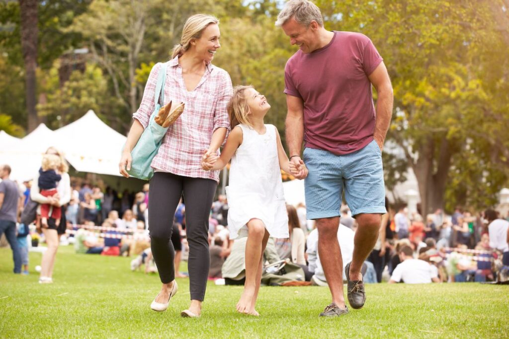 A mother and father holding hands with their barefoot daughter between them, walking on grass at an outdoor fair with white tents and a crowd blurred behind them.