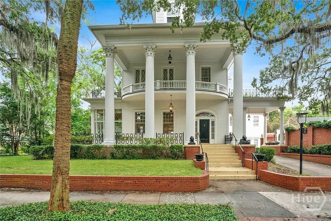 Historic white mansion with large columns, wraparound porch, and manicured lawn under a clear blue sky.
