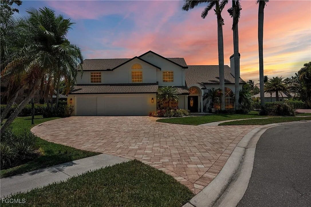 Two-story house with a large driveway, surrounded by palm trees, at sunset.