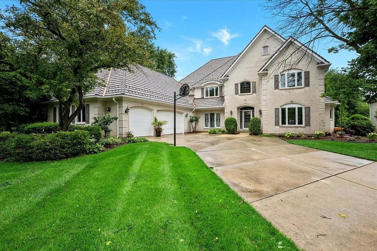 Large two-story house with a manicured lawn, trees, and a spacious driveway on a clear day.