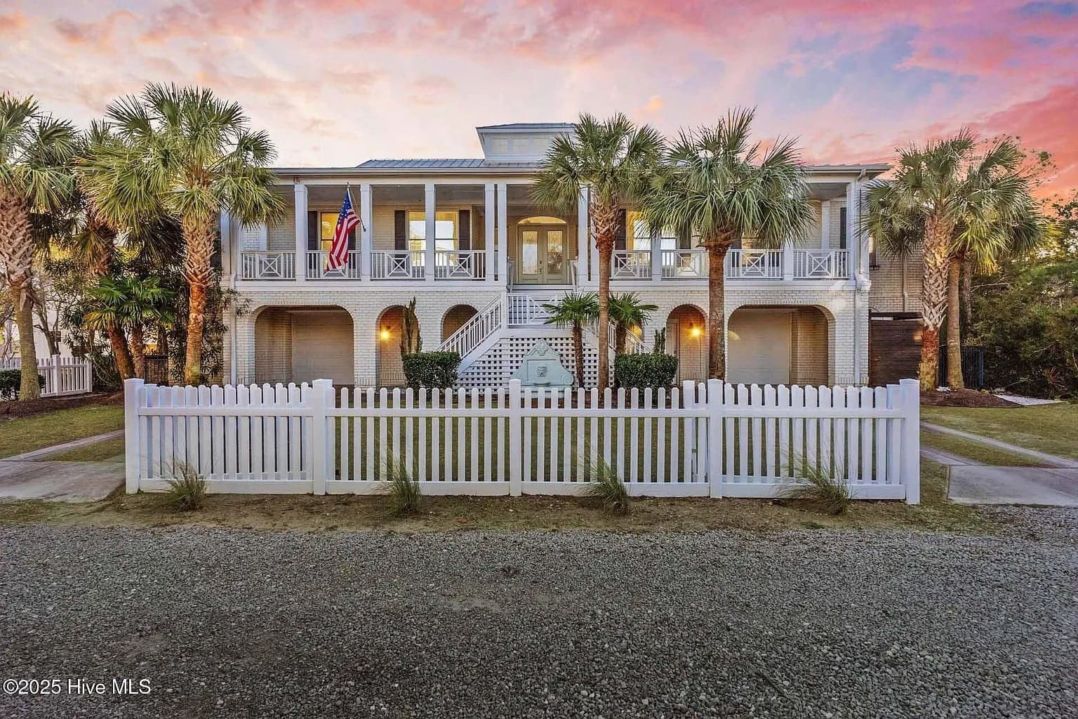Two-story house with palm trees, white fence, and American flag at sunset.