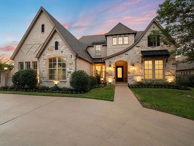 Large stone house with gabled roof, illuminated windows, and a wide driveway at sunset.