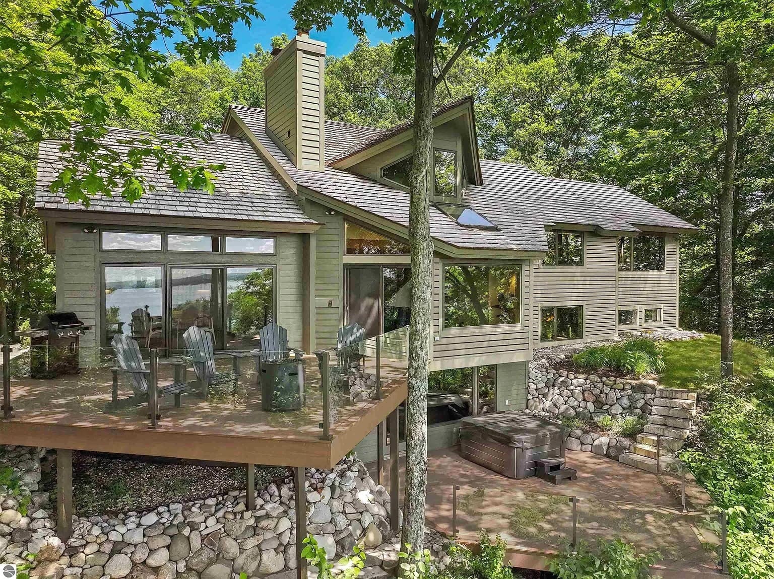 Modern lakeside house with large windows, wooden deck, outdoor seating, and surrounded by trees.