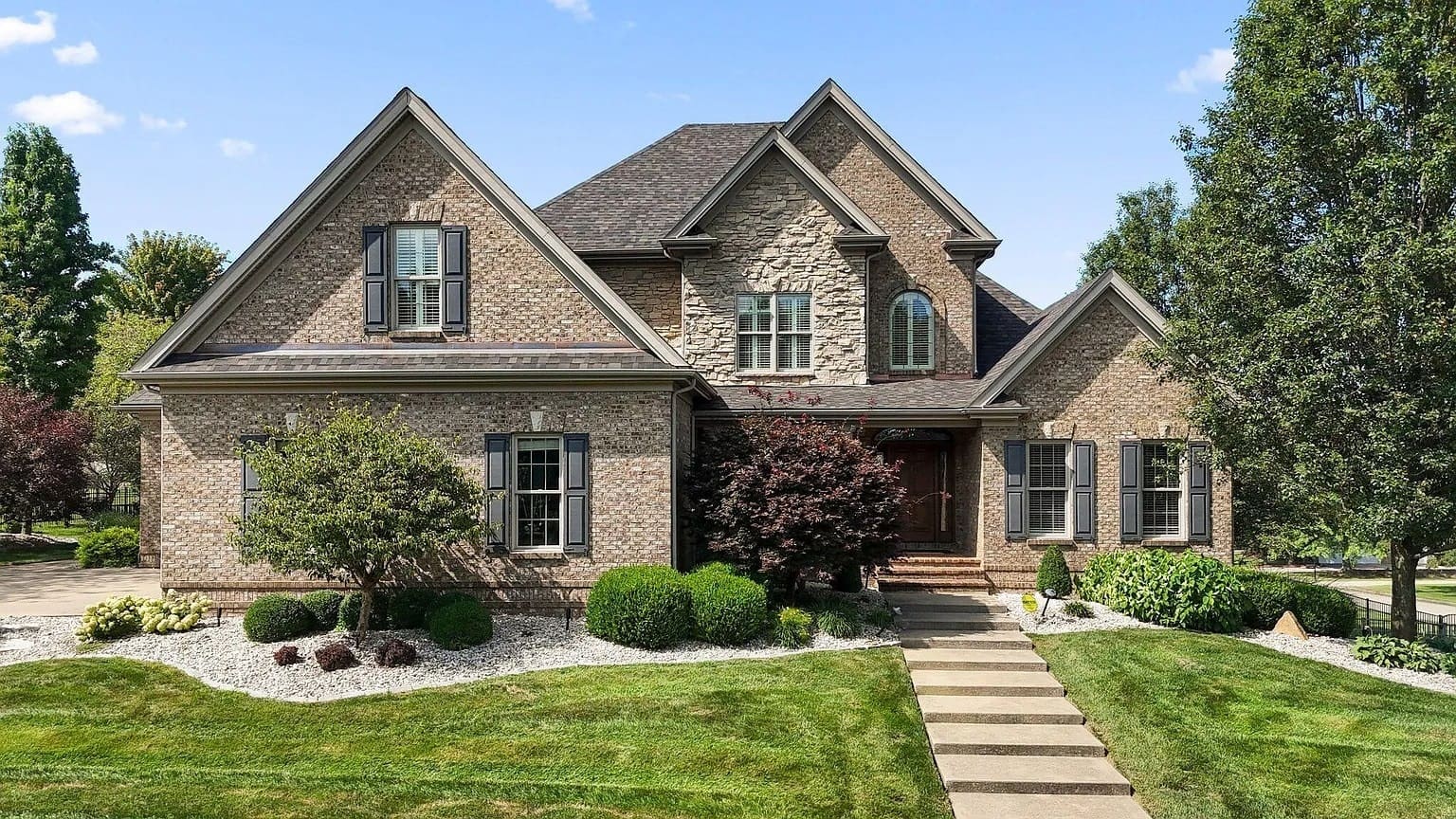 Two-story brick house with landscaped front yard, trees, and a stone walkway under a clear sky.