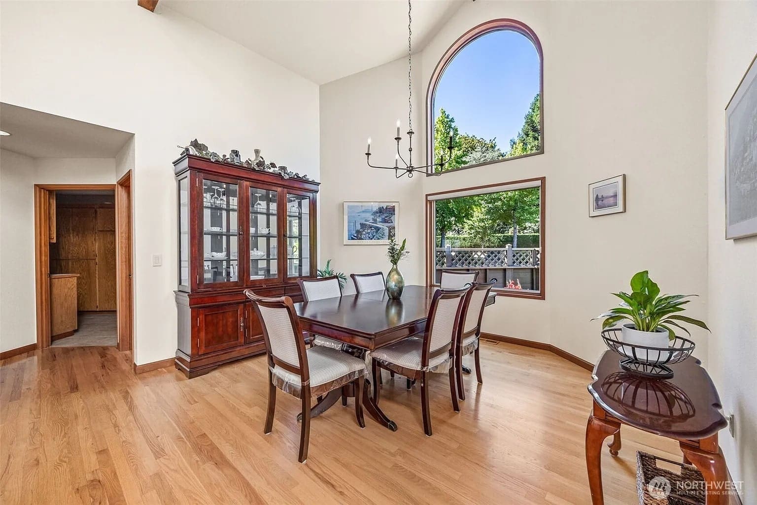 Dining room with a wooden table, chairs, china cabinet, large arched window, and plants.