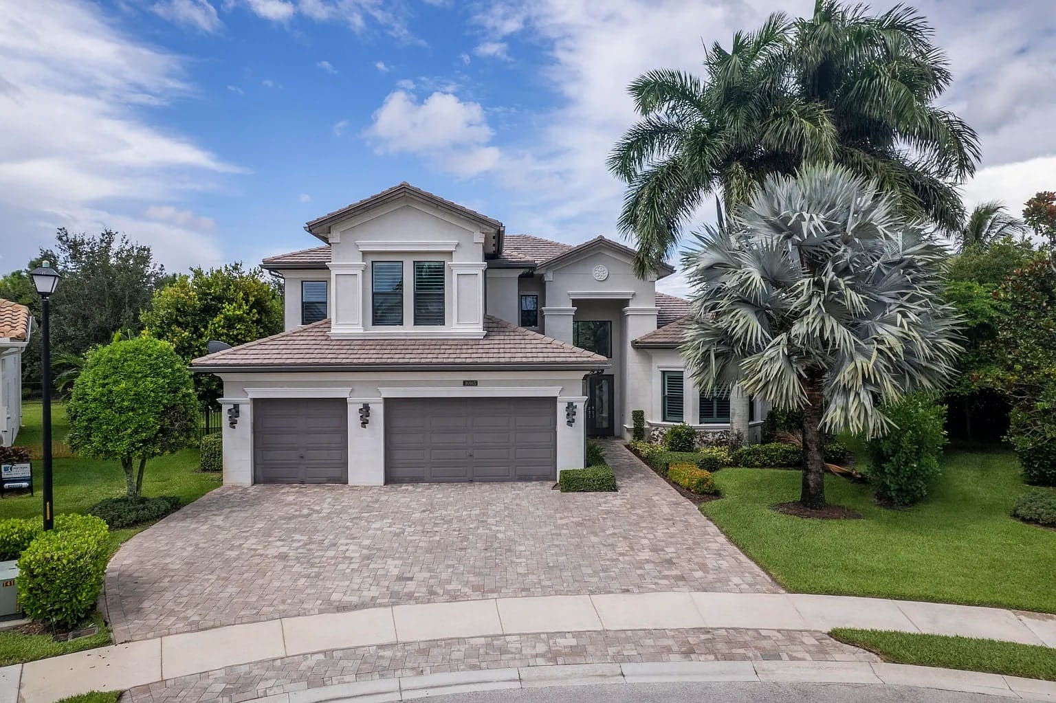 Two-story modern house with three-car garage, palm trees, and a paved driveway under a blue sky.
