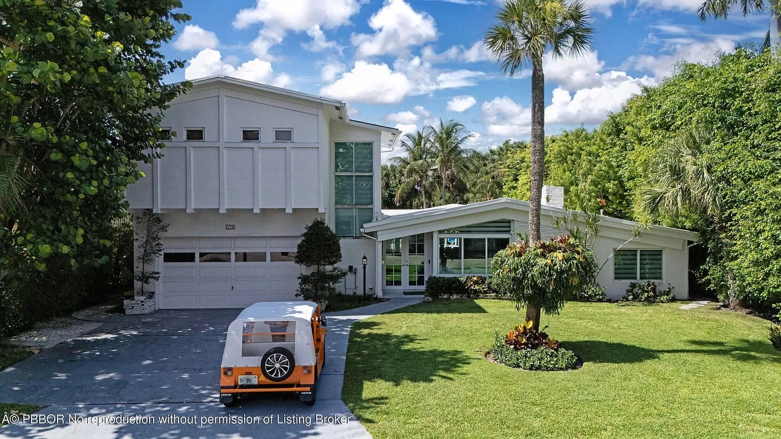 Modern two-story house with large windows, surrounded by lush greenery, and a small orange vehicle.
