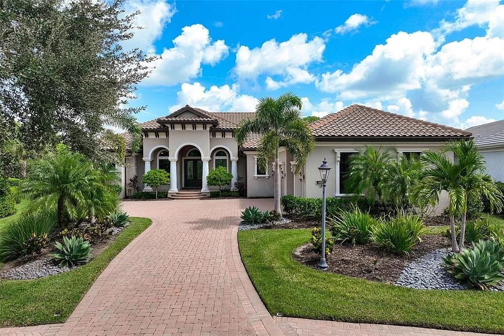 Single-story Mediterranean-style house with arched entry, palm trees, and brick driveway under a blue sky.
