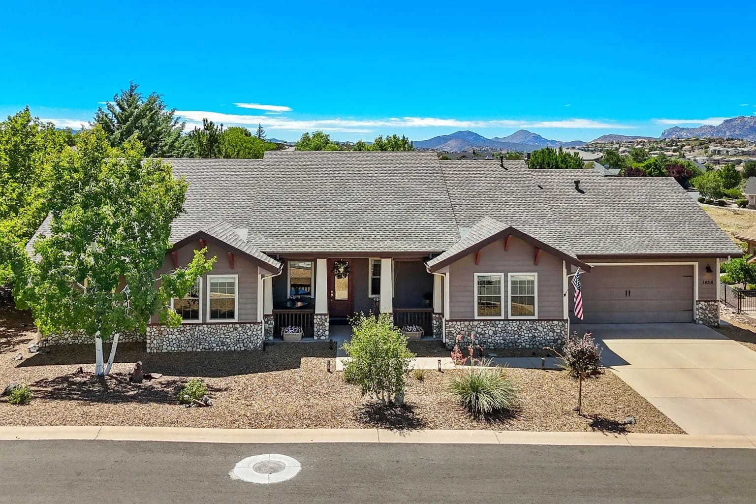 Single-story house with a stone facade, surrounded by trees, mountains in the background, clear sky.