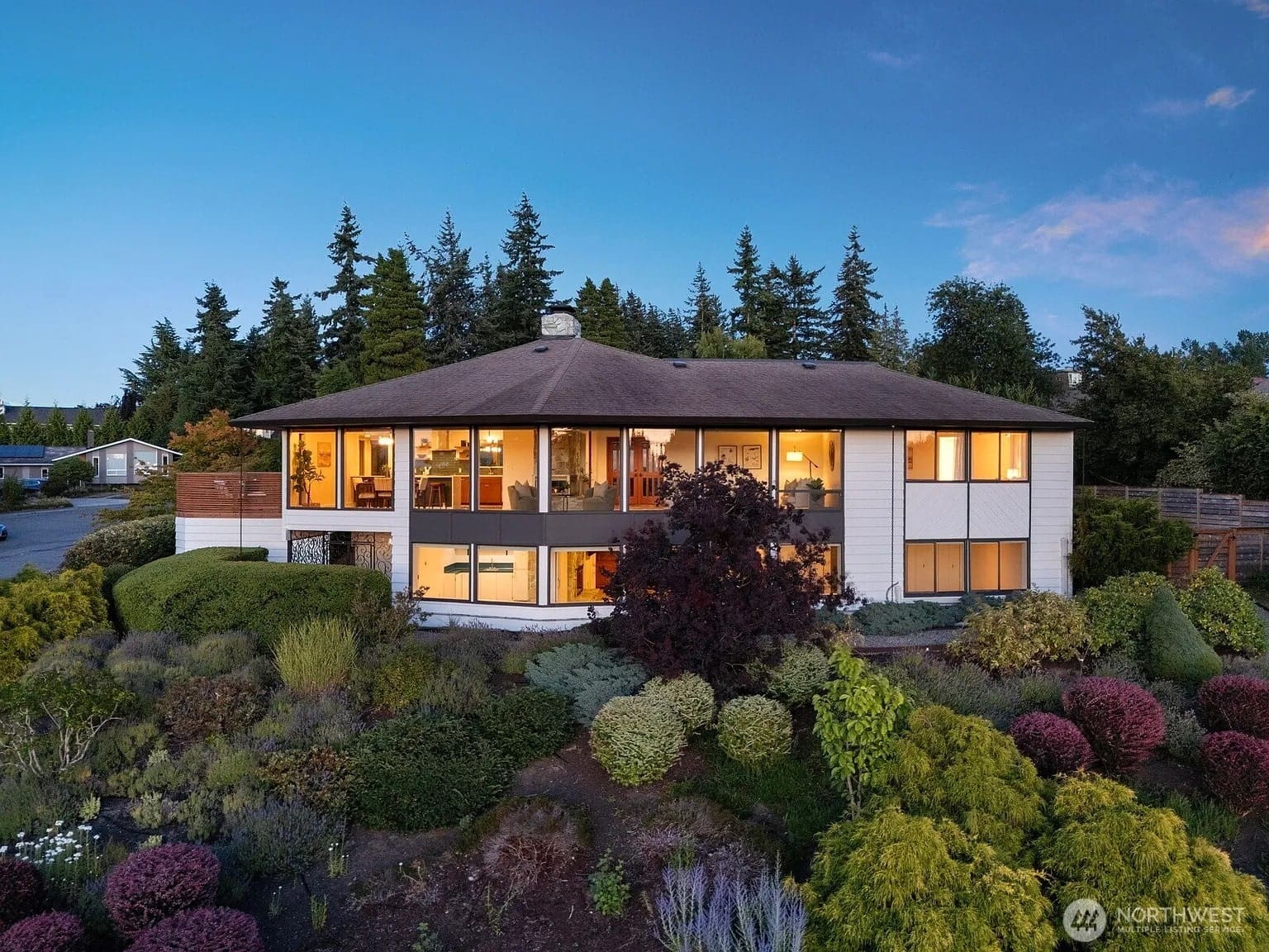 Two-story house with large windows, surrounded by lush greenery and trees, at dusk.