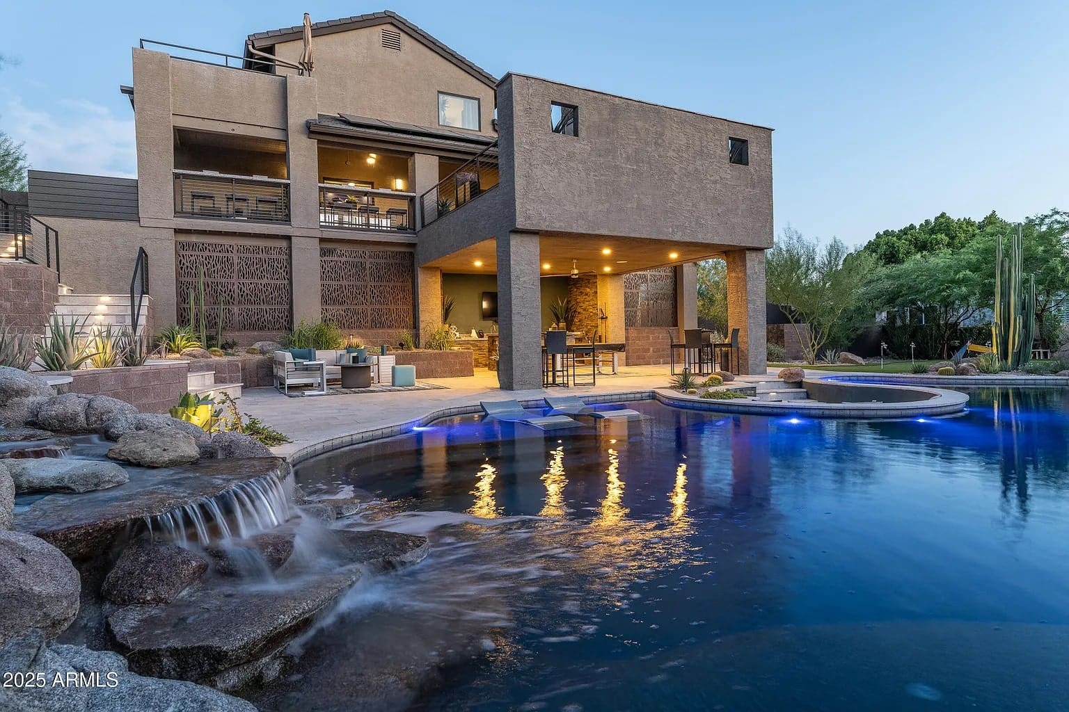 Modern house with large patio, pool, waterfall, and outdoor seating area at dusk.