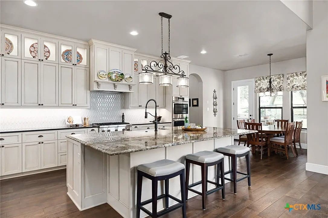 Spacious kitchen with white cabinets, large granite island, pendant lighting, and dining area with windows.