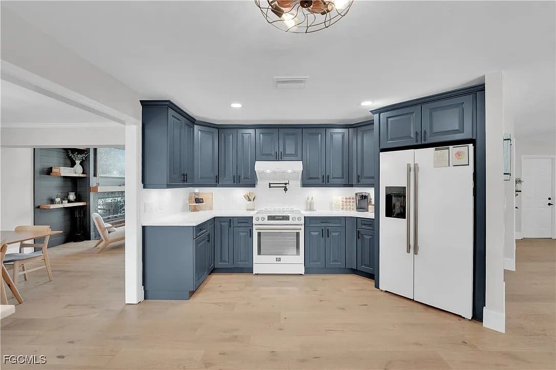 Modern kitchen with blue cabinets, white appliances, and light wood flooring. Dining area visible in background.