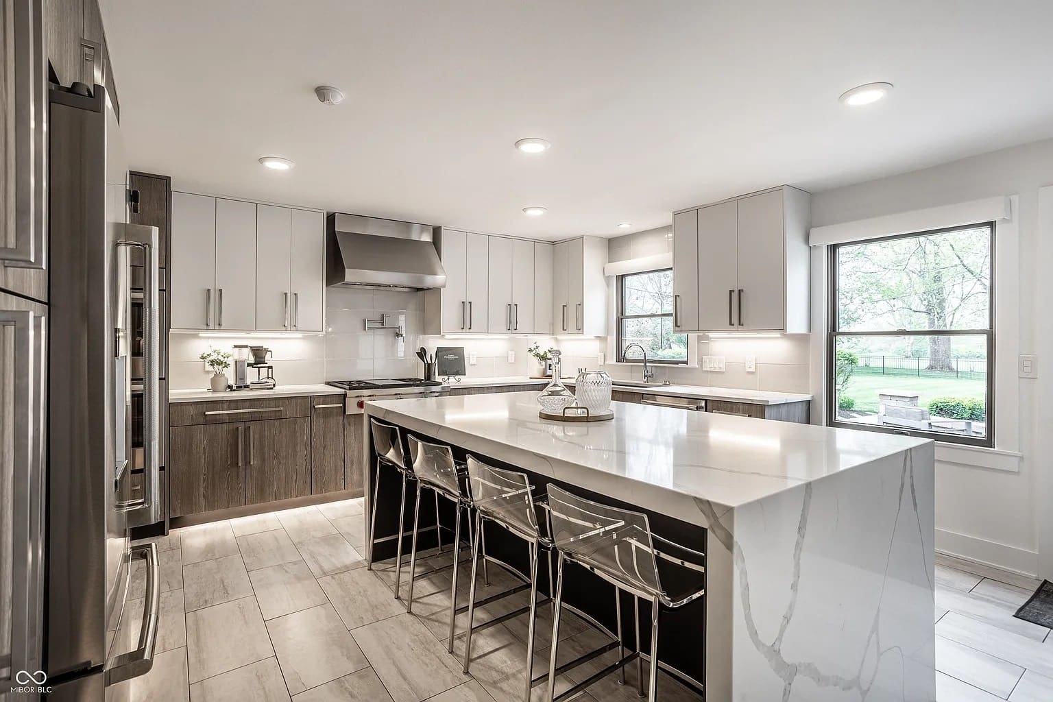 Modern kitchen with marble island, bar stools, stainless steel appliances, and large windows overlooking greenery.