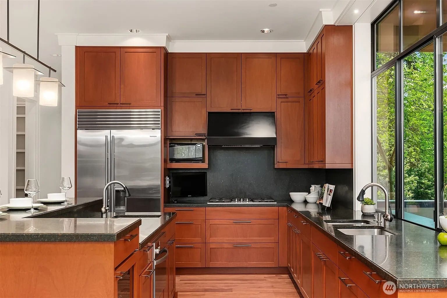 Modern kitchen with wooden cabinets, stainless steel appliances, and large windows overlooking greenery.