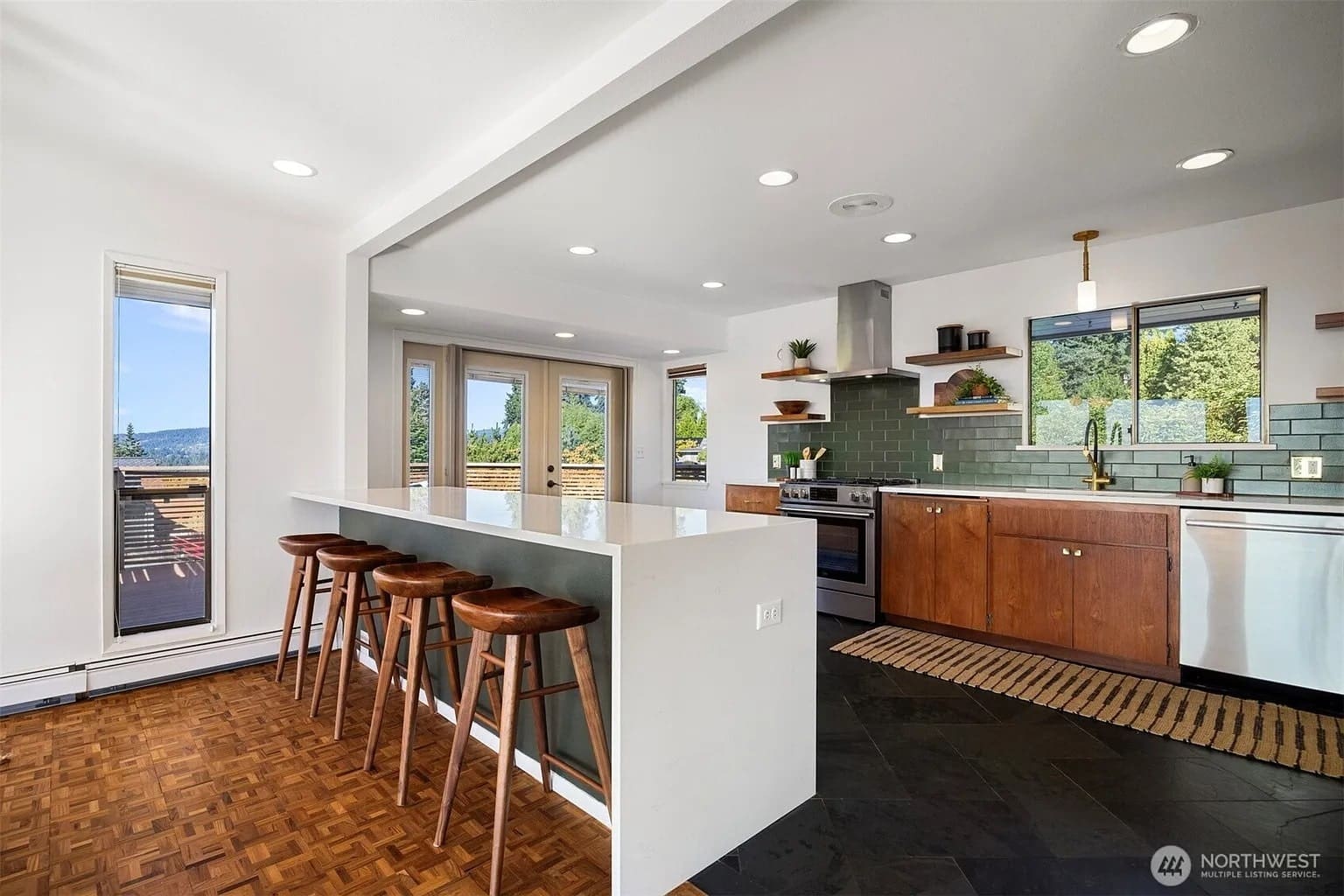 Modern kitchen with white island, wooden stools, green tile backsplash, and large windows with scenic view.