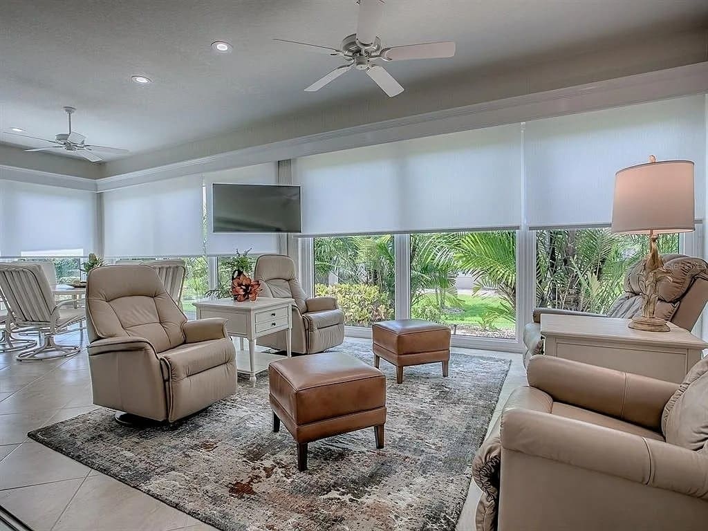 Bright living room with recliners, ottomans, large windows, and a view of greenery outside.
