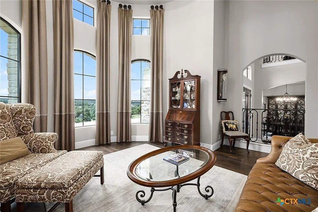 Elegant living room with tall windows, patterned armchairs, a glass coffee table, and a wooden cabinet.