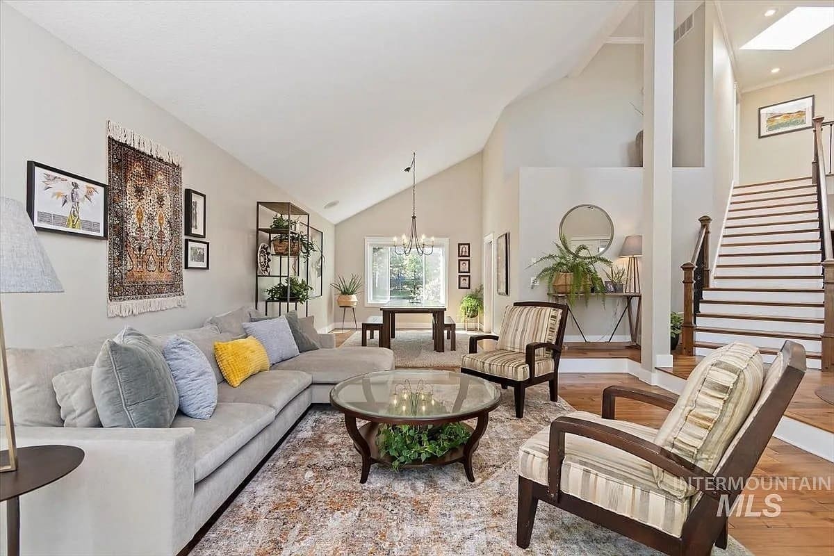 Spacious living room with beige sofa, patterned chairs, glass coffee table, and staircase. Bright and airy.