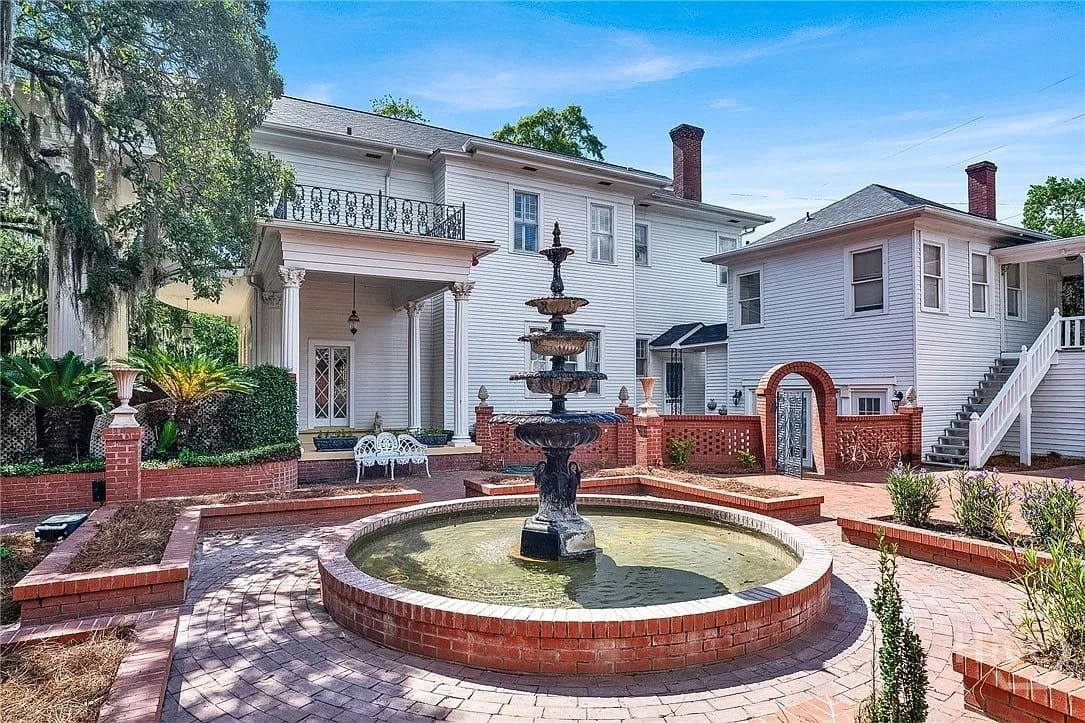 Historic white house with a large brick courtyard, central fountain, and surrounding greenery.
