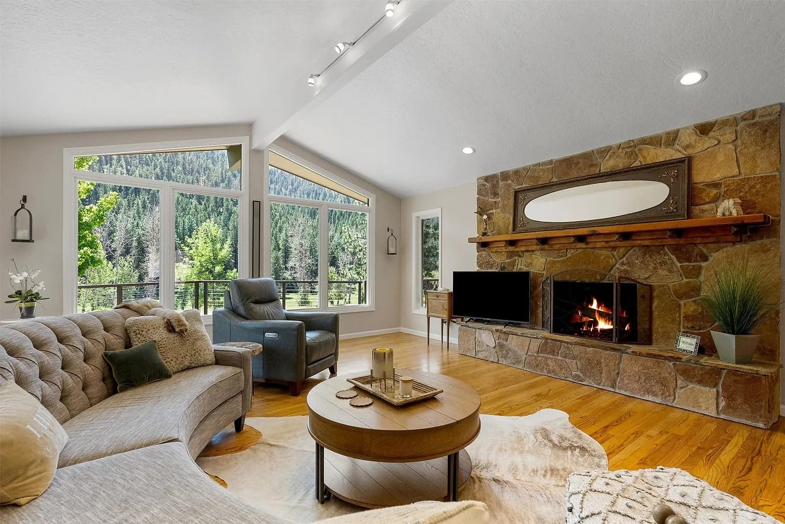 Cozy living room with a stone fireplace, large windows, and comfortable seating overlooking a forest view.