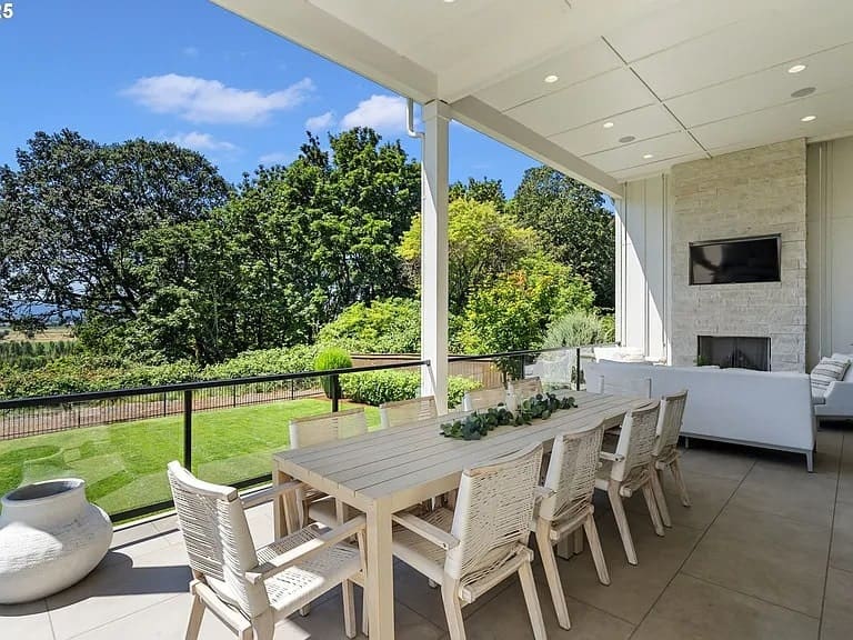 Outdoor patio with a long dining table, chairs, and a view of a garden and trees.