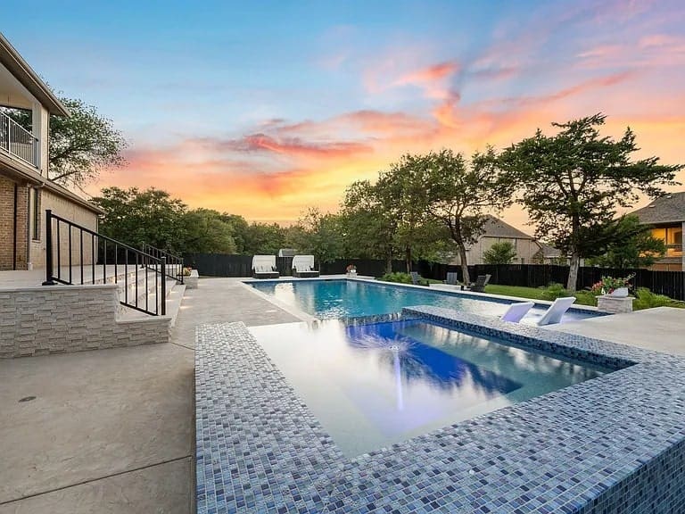 Backyard with a pool and hot tub at sunset, surrounded by trees and a patio area.