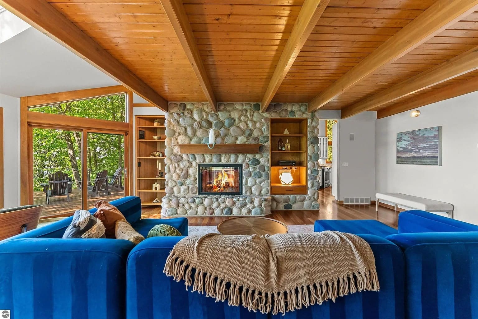 Living room with blue sofas, stone fireplace, wooden ceiling, and large windows overlooking greenery.