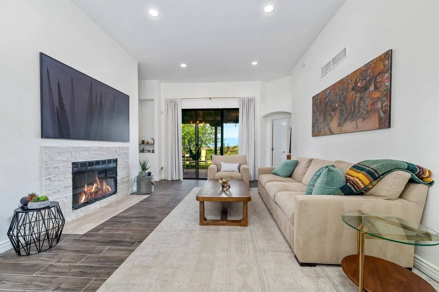 Modern living room with a fireplace, beige sofa, large artwork, and glass doors opening to a garden.