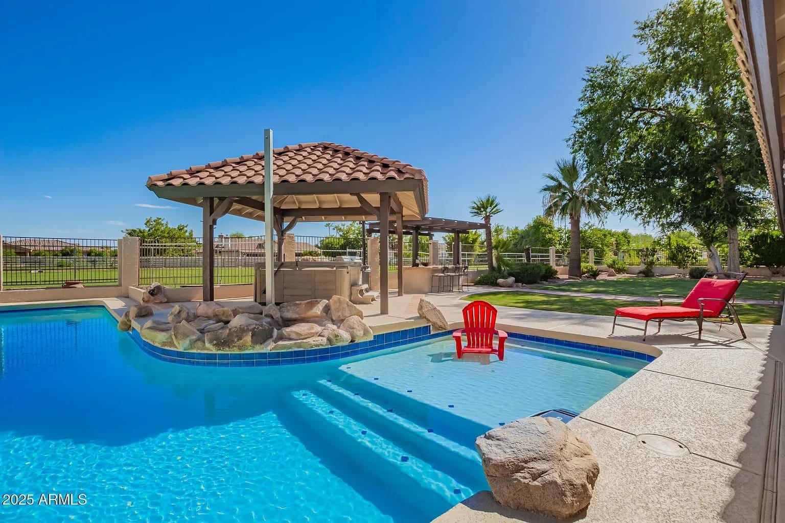 Outdoor pool with a gazebo, red chair, and lounge chair, surrounded by trees and a fence.
