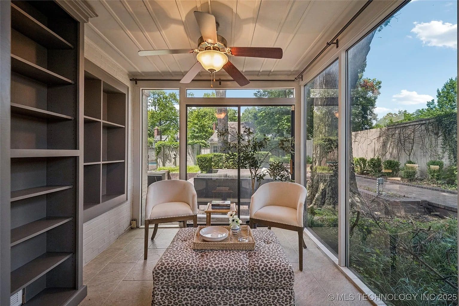 Sunroom with two chairs, a patterned ottoman, ceiling fan, and large windows overlooking a garden.