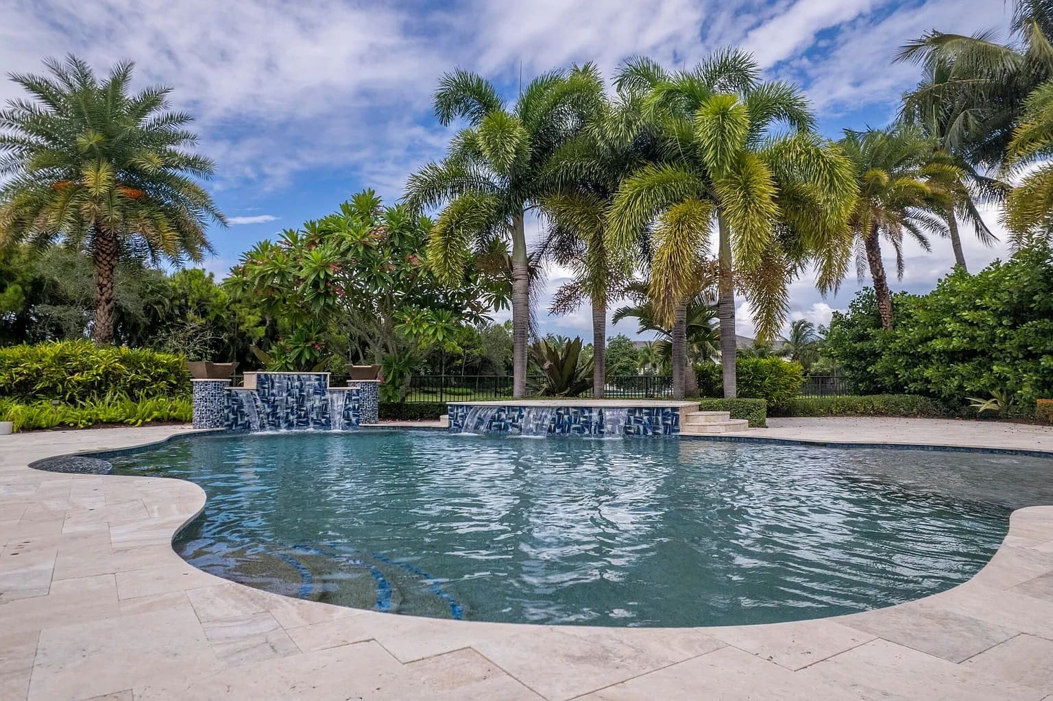 Outdoor pool with waterfall feature, surrounded by palm trees and lush greenery under a blue sky.