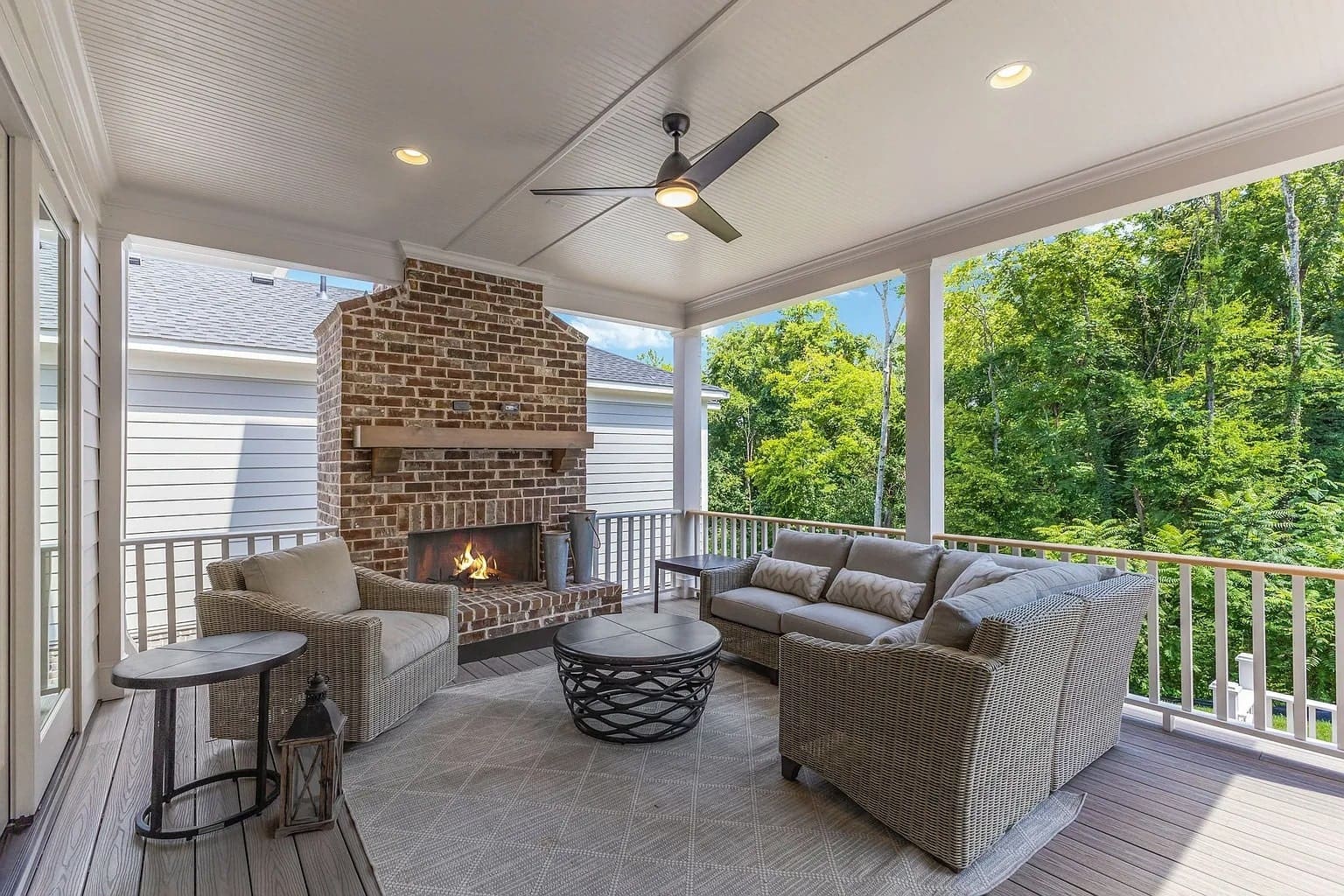 Covered patio with brick fireplace, wicker furniture, ceiling fan, and view of lush greenery.