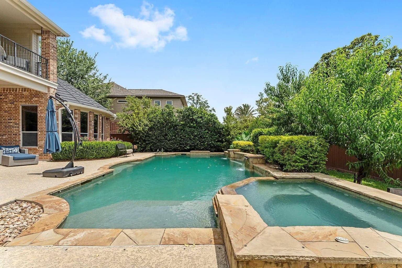 Backyard with a swimming pool, hot tub, and surrounding greenery on a sunny day.