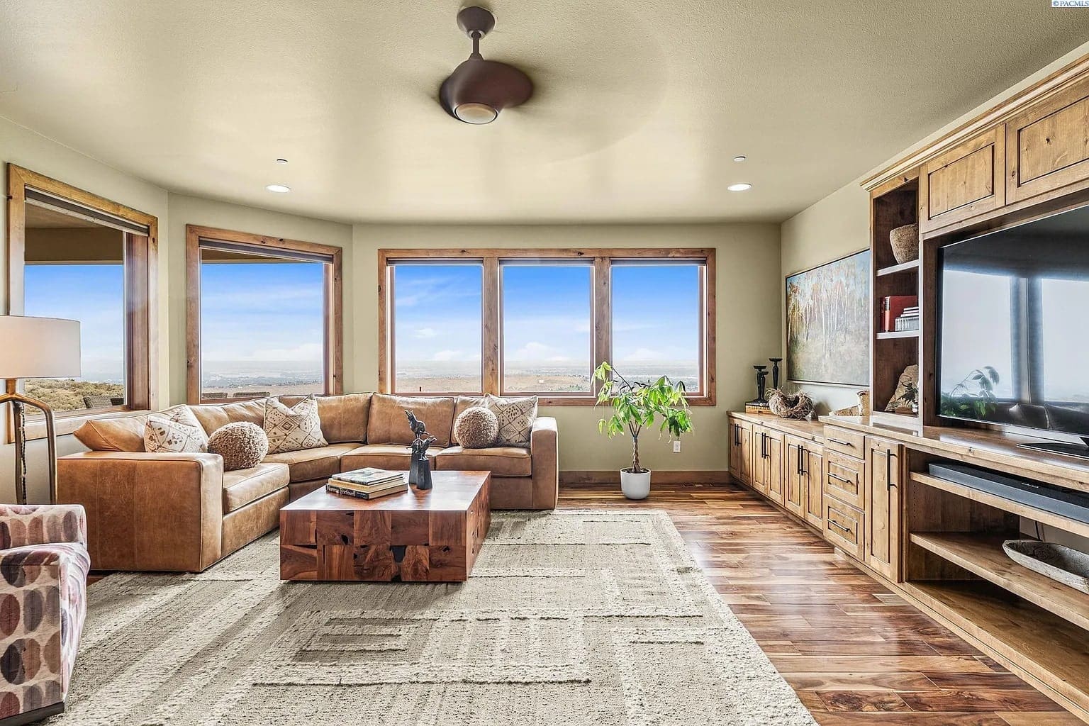 Spacious living room with large windows, brown sectional sofa, wooden furniture, and a ceiling fan.