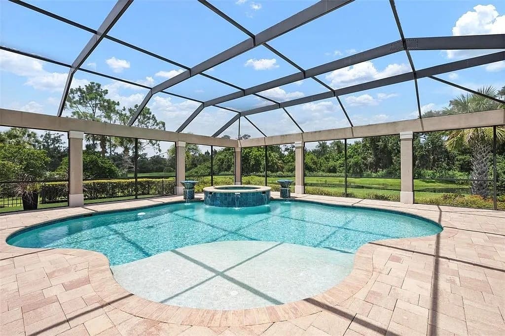 Enclosed outdoor pool with a central hot tub, surrounded by greenery and a clear blue sky.