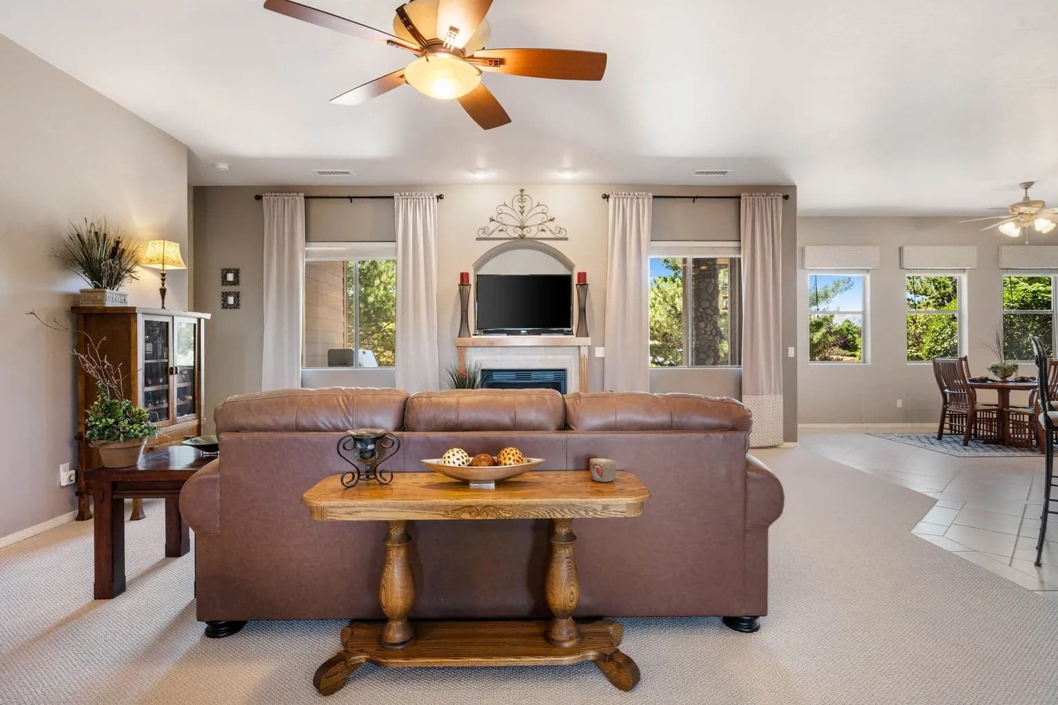 Living room with brown sofa, ceiling fan, TV, and large windows overlooking greenery.