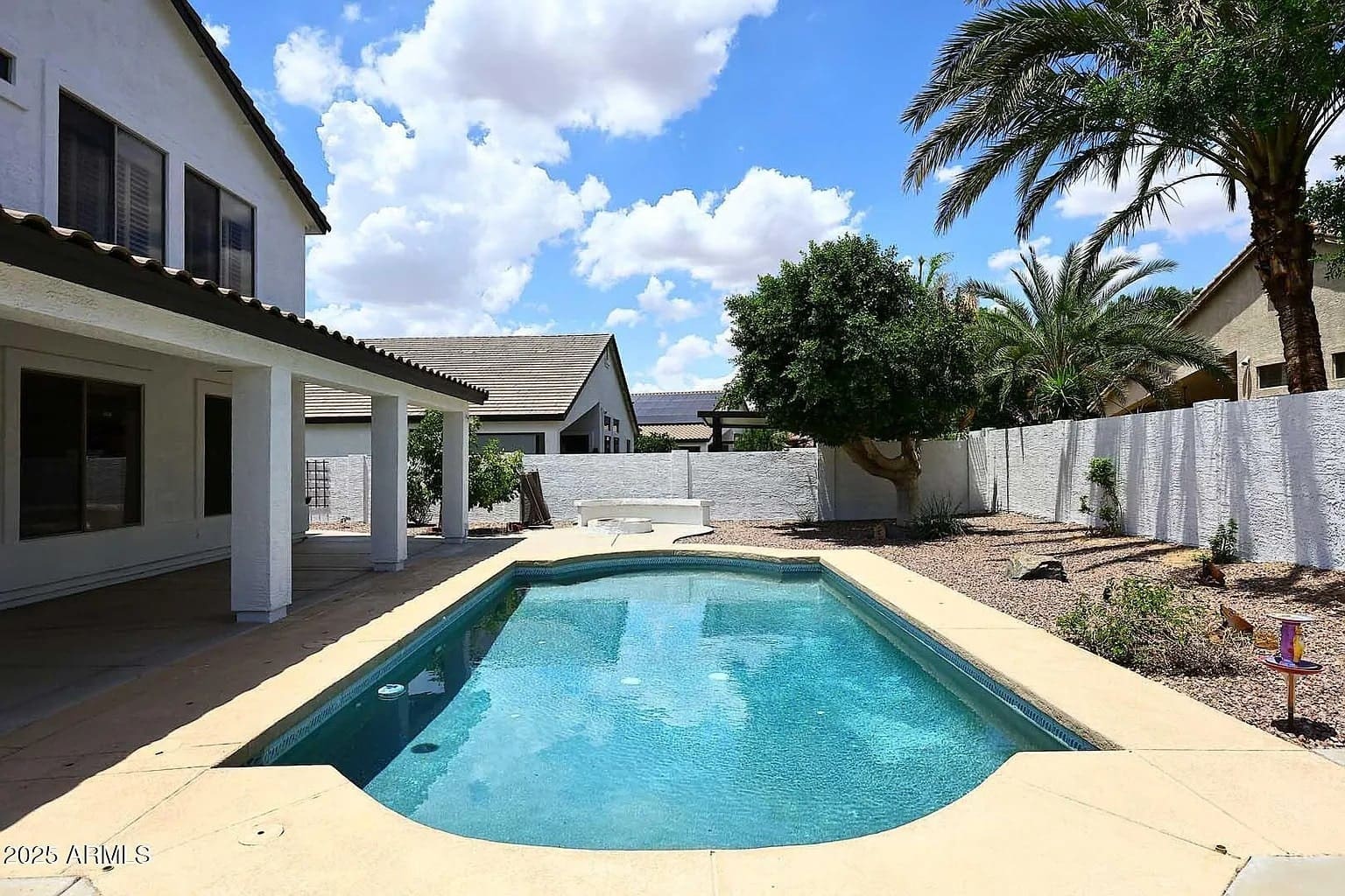 Backyard with a swimming pool, surrounded by a white fence, palm trees, and a covered patio.